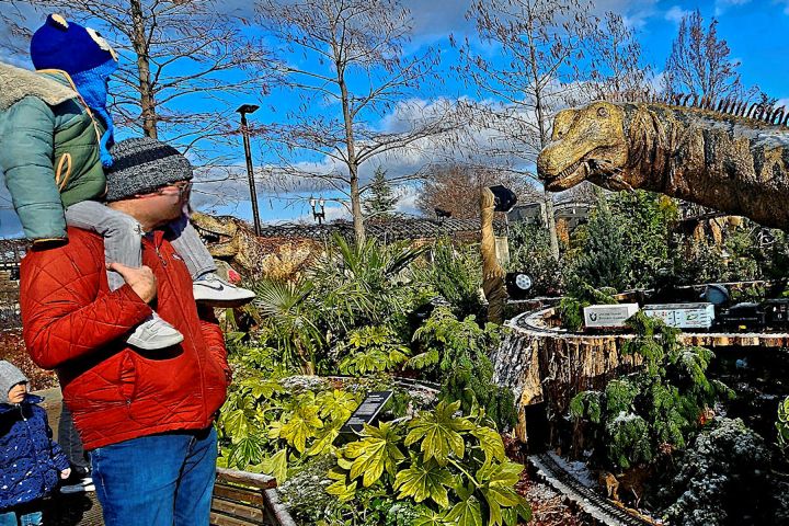 Man holding child on shoulders looks at dinosaur model in outdoor display with train at the U.S. Botanic Garden.
