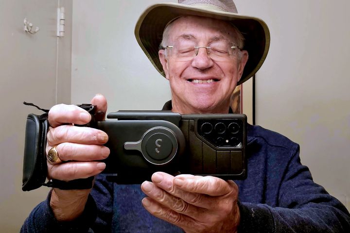Man in a hat smiling, taking a mirror selfie with a large camera phone.