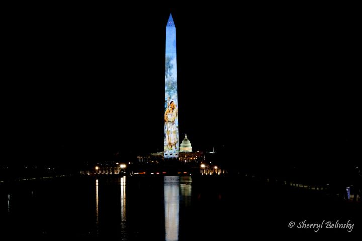 Washington Monument with Freedom250 illuminated art projection at night, Capitol building in background.