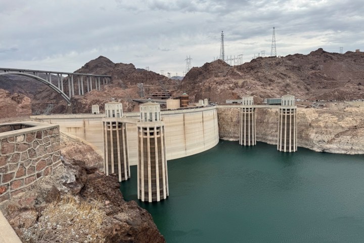 View of a large dam with spillways, surrounded by rocky landscape under cloudy skies.