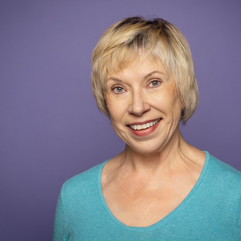 Smiling elderly woman with short blonde hair against a purple background.
